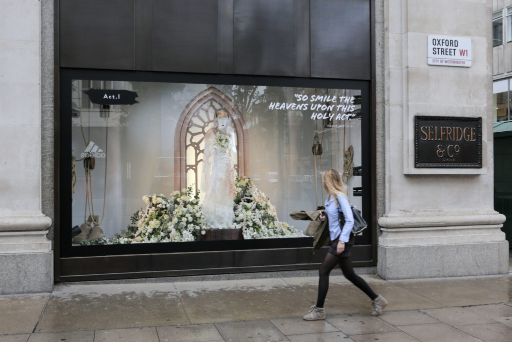 Selfridges window on Oxford Street dressed as a Shakespeare tableau, with a figure in white robes inside a gothic arch banked with flowers and a quotation across the glass, as a passer-by walks along the pavement.