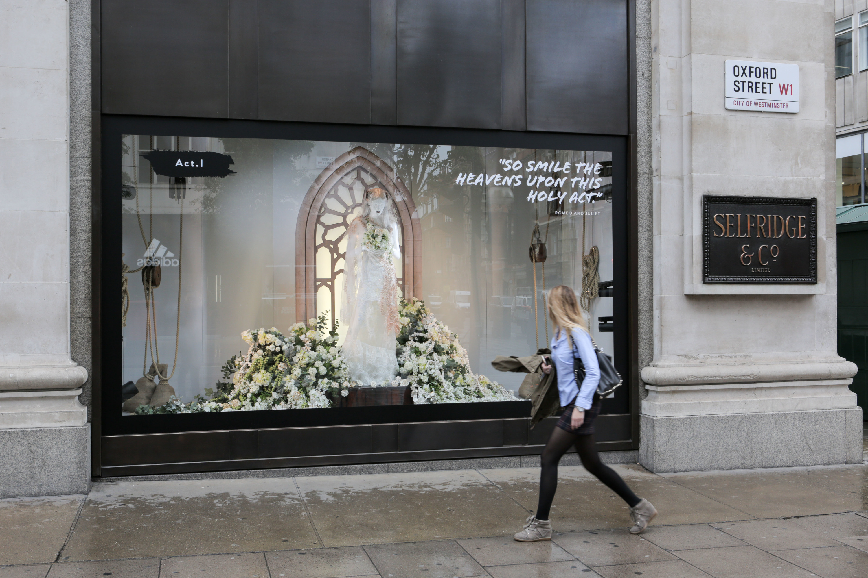 A large Selfridges window display on Oxford Street beneath the Oxford Street W1 street sign, with a passer-by walking along the pavement.