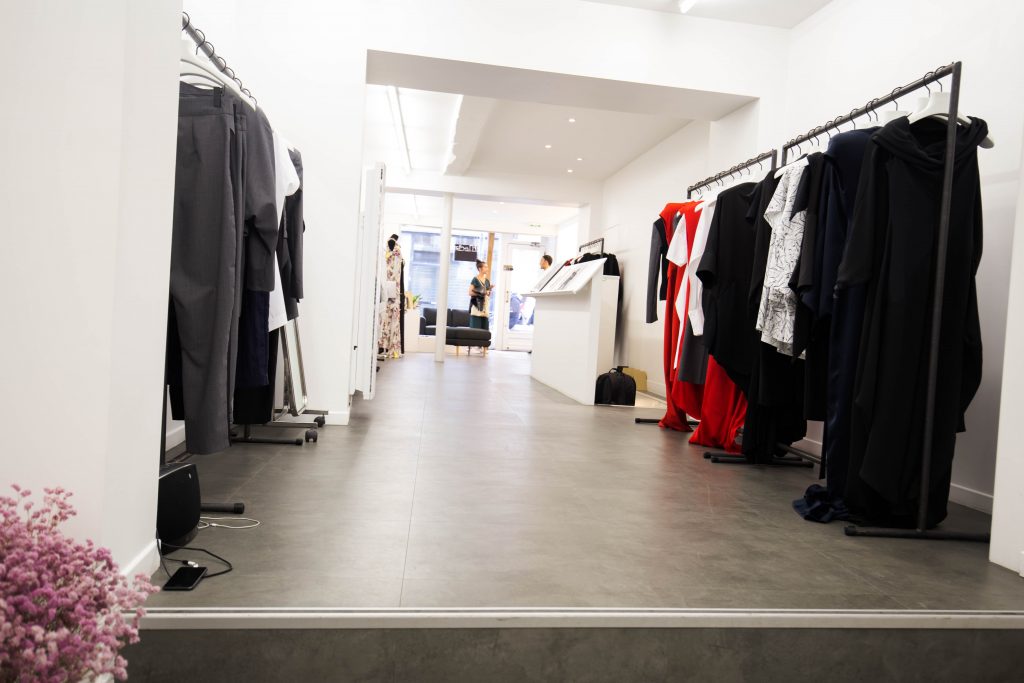 Narrow showroom with a polished screed floor, rails of black, red and printed draped garments along both walls, a raised white platform and glazed doors to the street at the far end.