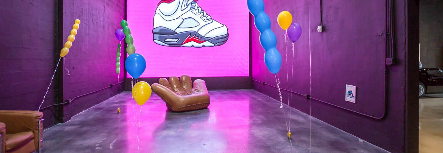 Pop-up room with purple and pink painted brick walls, a large backlit screen showing a white and red high-top trainer, a tan hand-shaped armchair and helium balloons weighted to the floor.
