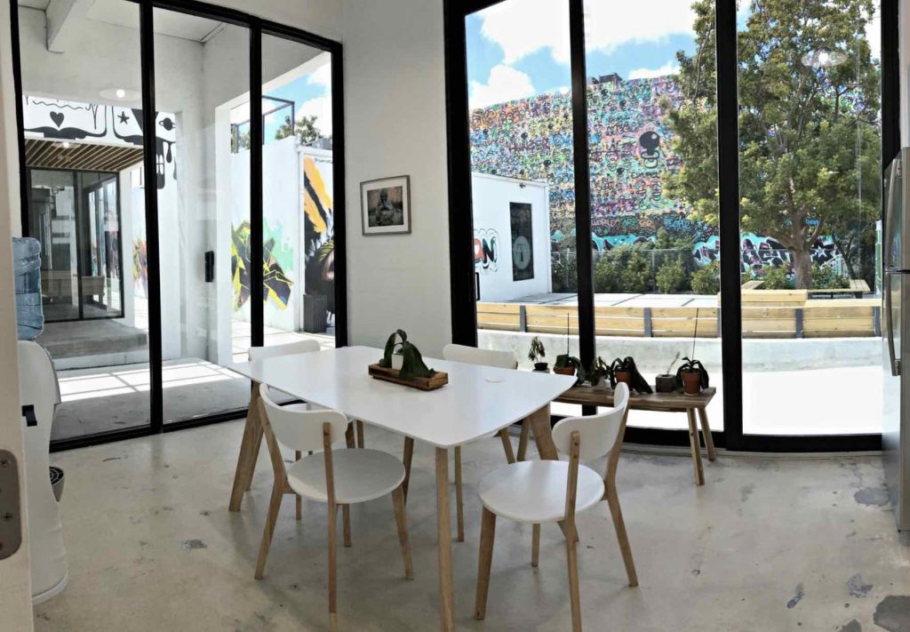 White space with a polished concrete floor and black steel sliding doors, a white table with pale timber chairs, looking out to a large wall covered edge to edge in dense graffiti and palms beyond.