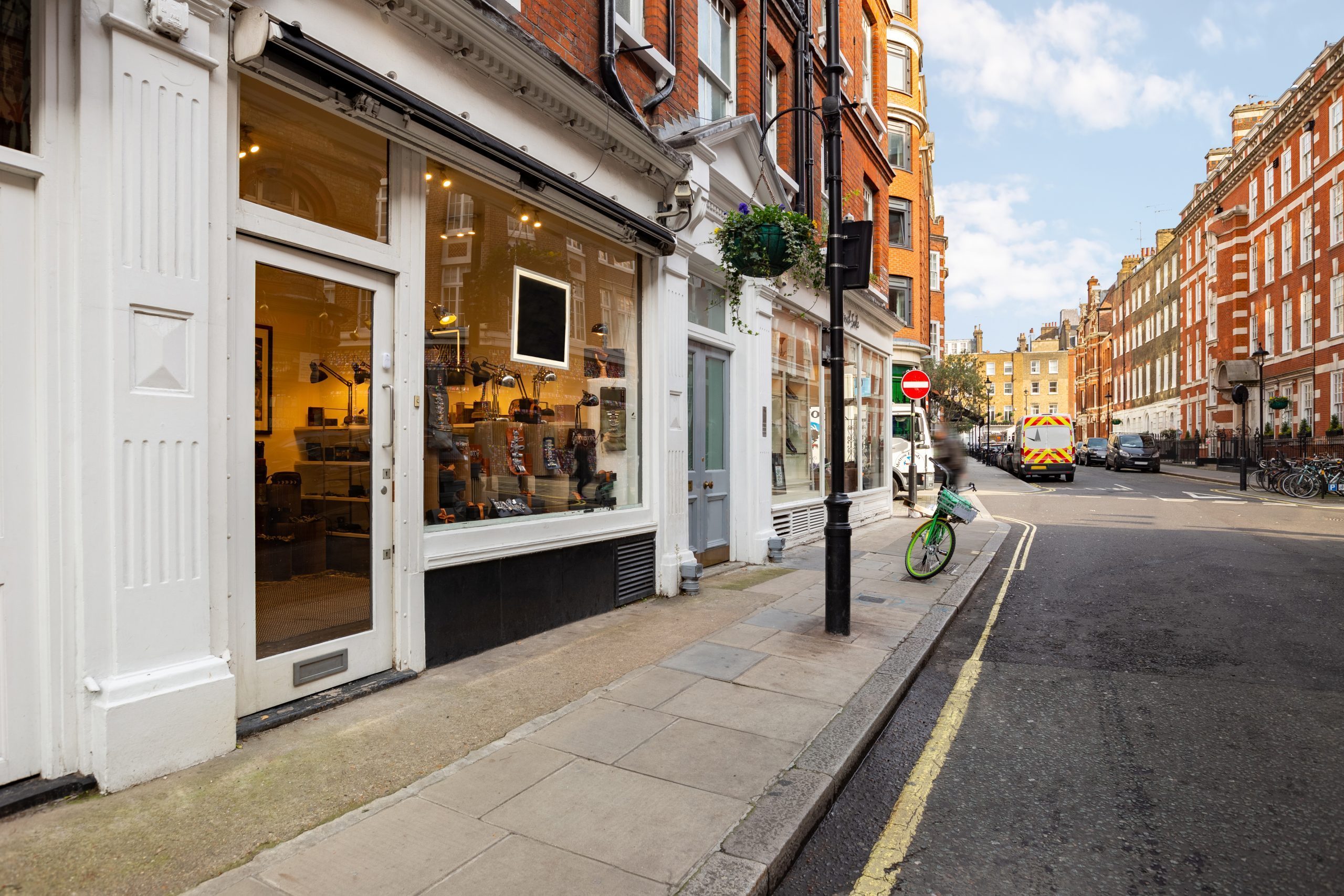 A white stucco Marylebone shopfront with a lit window display, on a narrow street lined with red-brick mansion blocks and a hanging basket above the pavement.