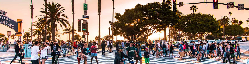Crowds and cyclists crossing Ocean Avenue at sunset in Santa Monica, with tall palms along the boulevard and the yacht harbor sign on the corner building.