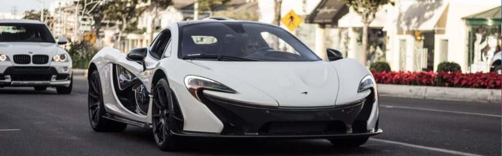A white McLaren supercar driving along a Beverly Hills street, with boutique frontage, clipped street trees and red seasonal planting behind it.