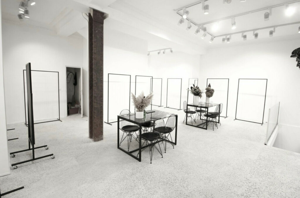 White showroom with a terrazzo floor, empty black steel garment rails and two small meeting tables.