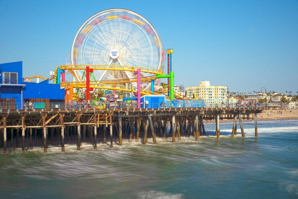 Santa Monica Pier seen from the water, with the Pacific Park ferris wheel and roller coaster on the deck and the beach running north behind it.