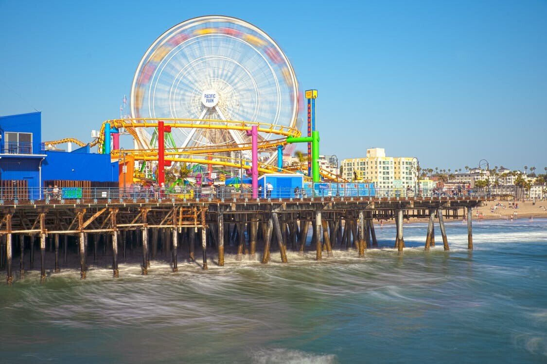 Santa Monica Pier seen from the water, with the Pacific Park ferris wheel and roller coaster on the deck, timber piles below and the beach and hotels running north behind it.
