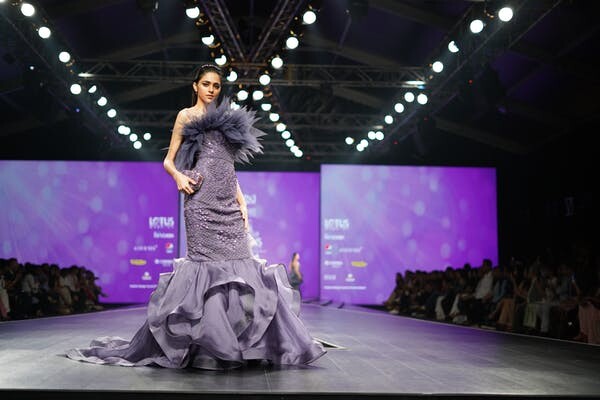 Mannequin en robe violette fluide sur le podium d'un défilé de la Fashion Week de Paris, sous les projecteurs.
