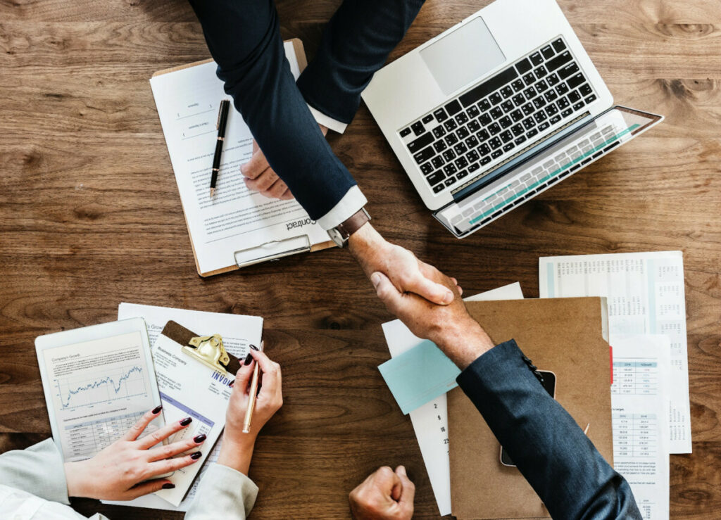 Overhead view of two people shaking hands across a timber desk spread with a clipboard contract, a laptop, invoices, printed charts and a pen, with a third person taking notes.