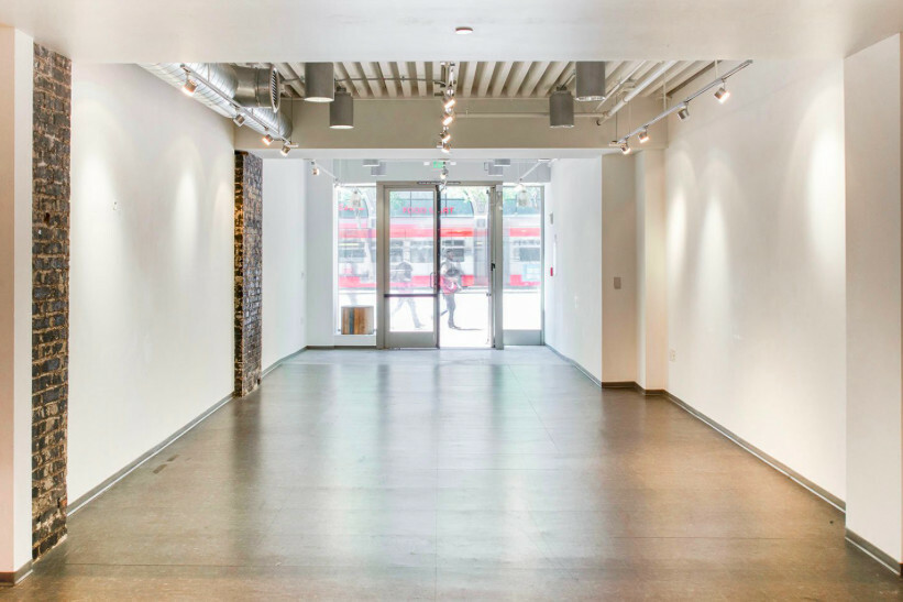 Long empty retail unit with white walls, an exposed dark brick pier, track lighting on an exposed ceiling and a glazed frontage onto a street where a white and red light rail vehicle is passing.