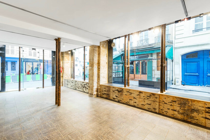 Empty retail unit with slim cast-iron columns, exposed limestone piers and brass-clad display plinths under the windows, glazed onto a street of cream stone buildings with wrought-iron balconies and a blue carriage door.