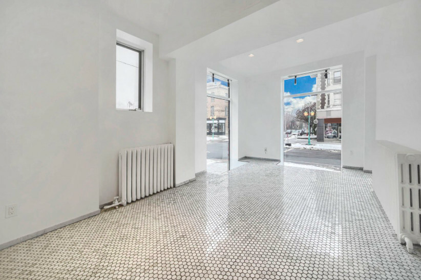 Empty white retail unit with a glossy penny tile floor, two cast-iron column radiators and a glazed frontage onto a snowy street.